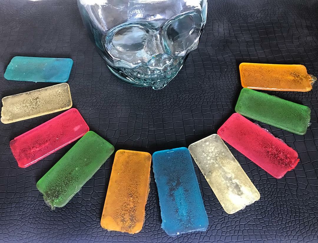 purAPPification, 2018 — arc of colored candy-cast mobile phones arranged around the glass skull vessel on a dark textured surface, detail view, by Hannes Bend