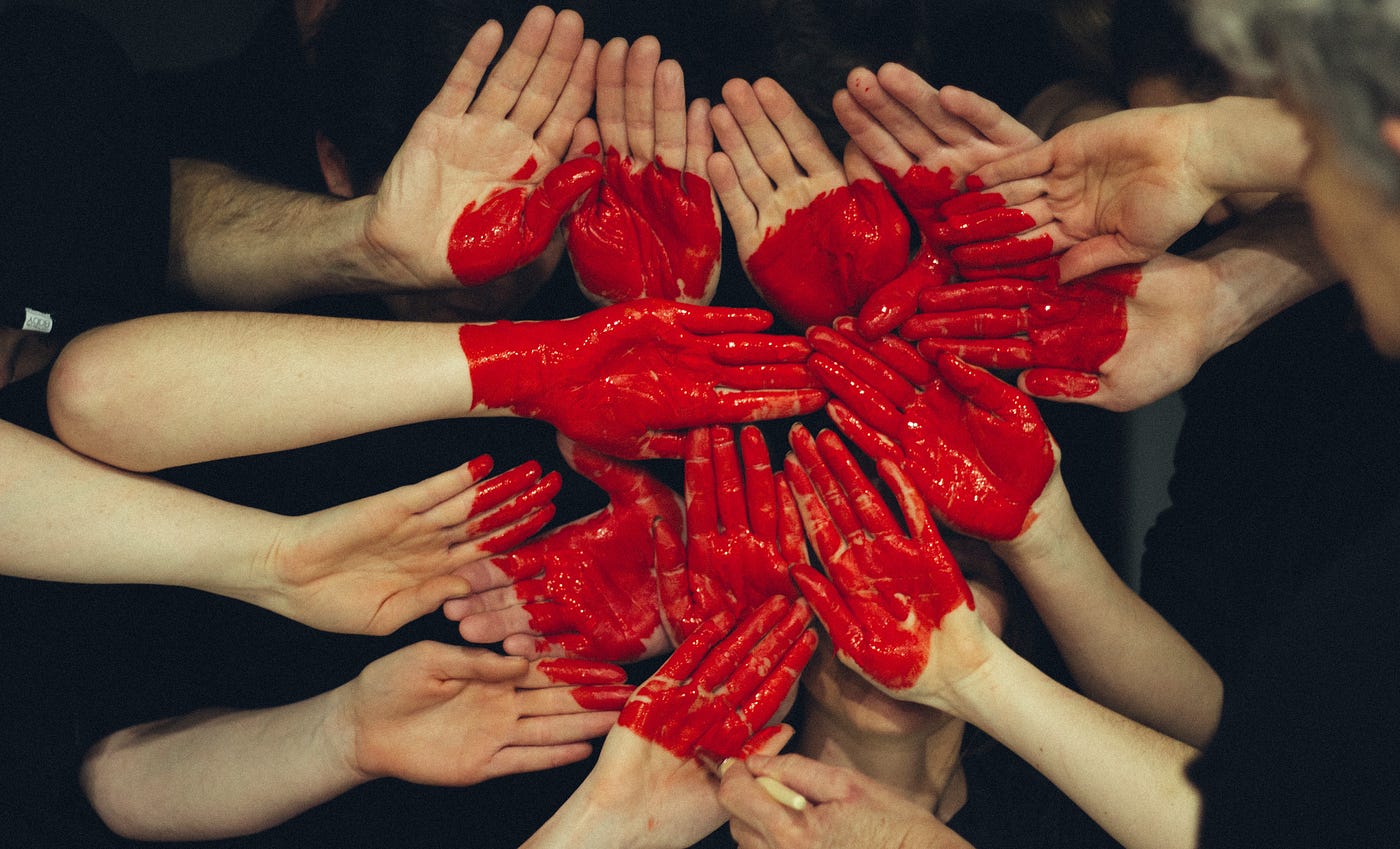 Many hands of different people, palms up and painted with red paint, arranged together so that the paint forms one large heart shape.