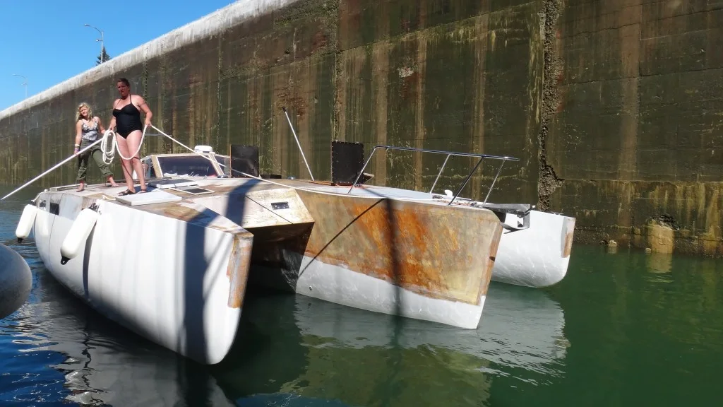 A white, partly rusted self-built trimaran moored against a high concrete lock wall, with two people handling lines on deck.