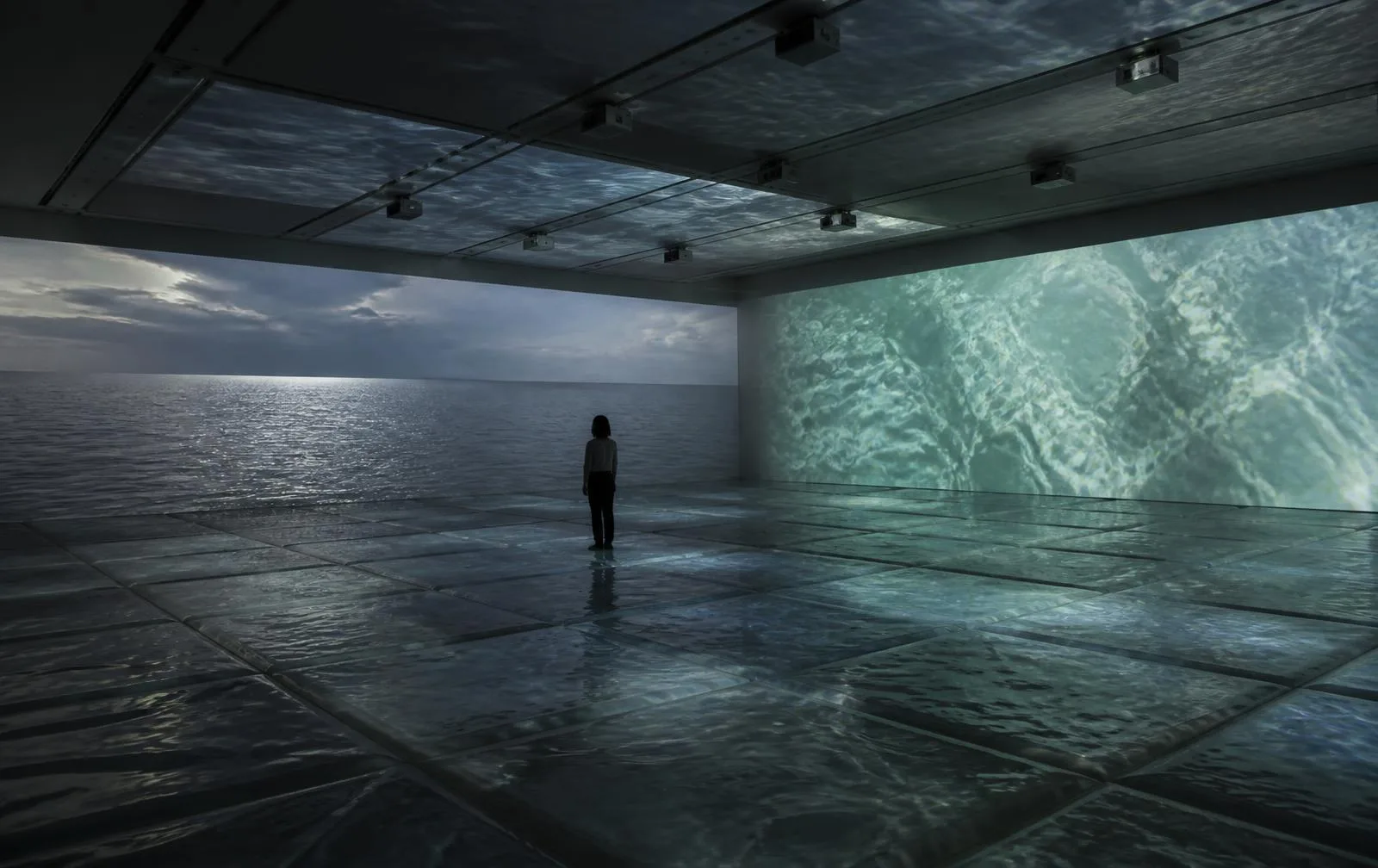 Interior view of Walking on VateR — a single visitor standing on the sealed water-filled modular floor between a wall carrying documentary footage of an ocean horizon and a wall carrying generated water, with imagery continuing across the ceiling