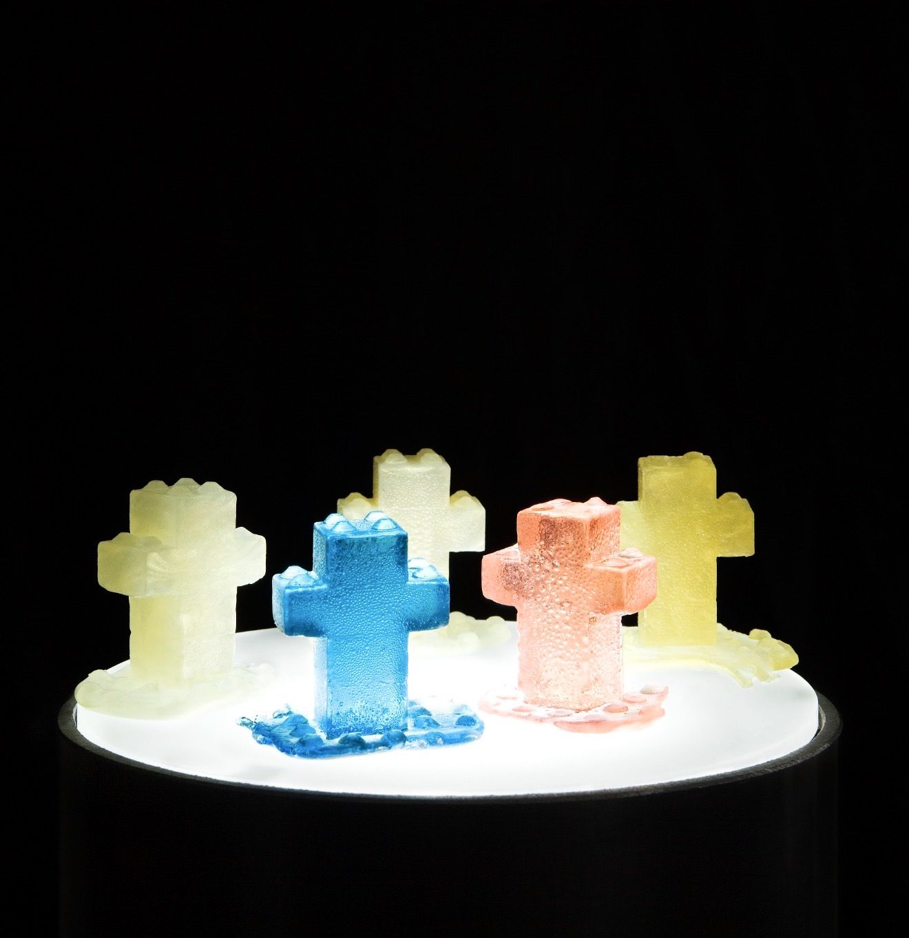 l'EGOs, 2011 — group of candy-cast crosses in yellow, blue, and pink at LEGO brick scale, illuminated on a pedestal, by Hannes Bend