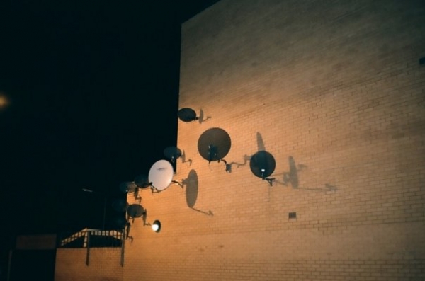 Satellites in the Night, 2010 — cluster of satellite dishes mounted on a brick façade at night, cast in warm streetlight, image related to the Satellites in the Night curatorial project at Freies Museum Berlin