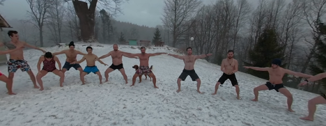 A group of people in shorts standing barefoot in snow in a horse stance during a winter breathing training session in a forest clearing.