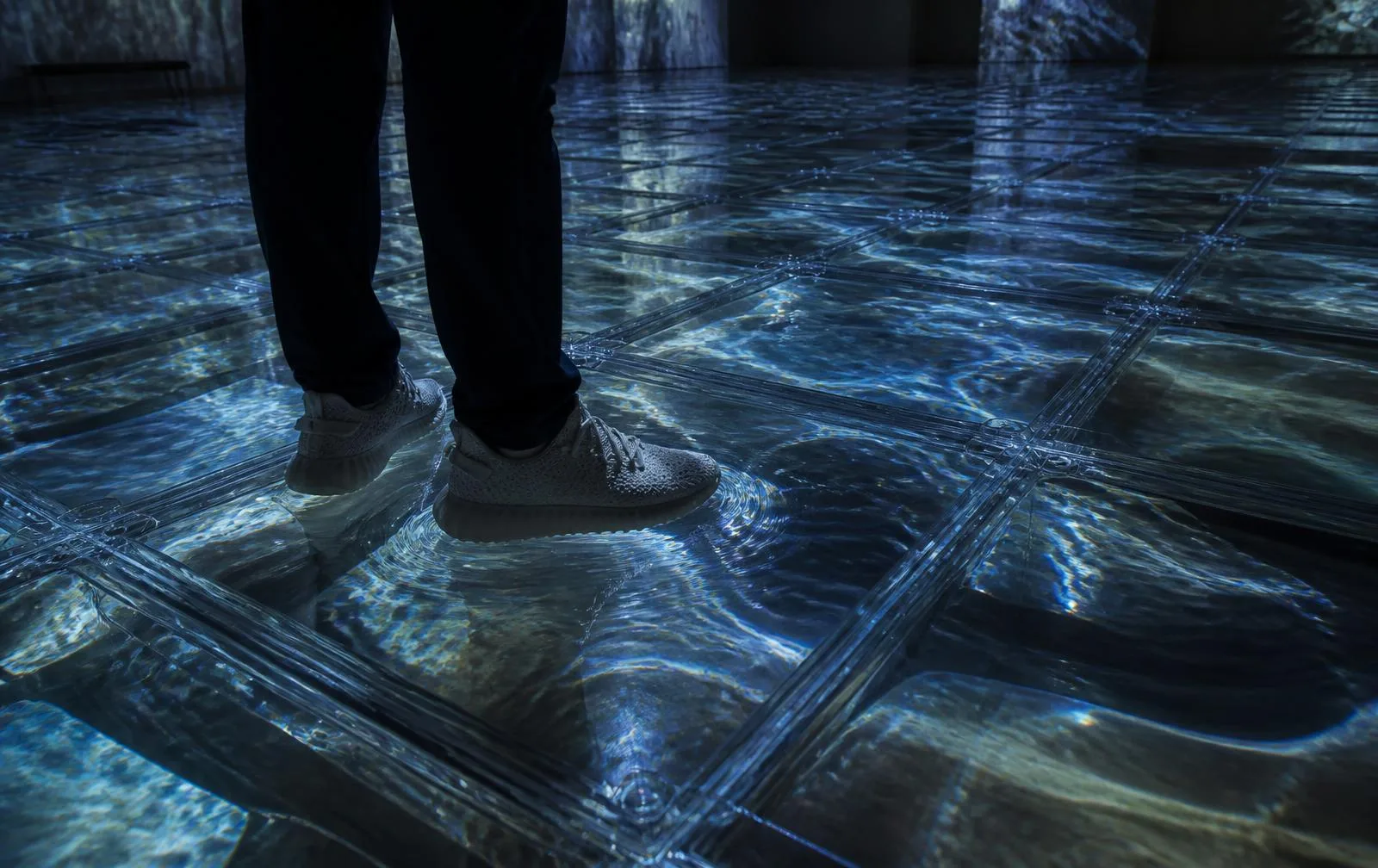 Floor detail of Walking on VateR — a visitor's feet on transparent sealed recycled-plastic cells filled with real water, the water displaced under body weight while projected water moves across the surface