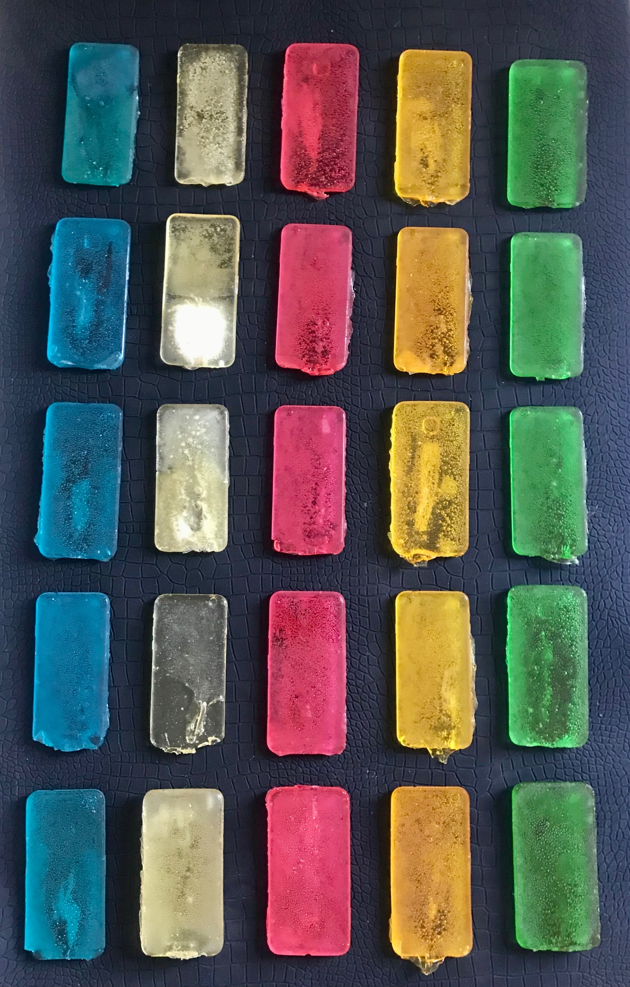 Sweet attention, 2018 — grid of twenty-five candy-cast mobile-phone forms in five colors (teal, clear, magenta, amber, green) arranged in a 5×5 grid on a black textured surface, by Hannes Bend