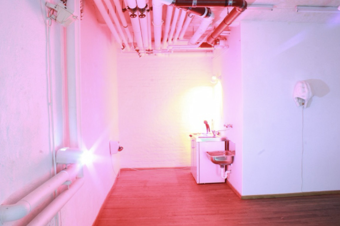 IN & OUT, 2004/2013 — warm pink-lit room with wall-mounted device, sink, and exposed pipes