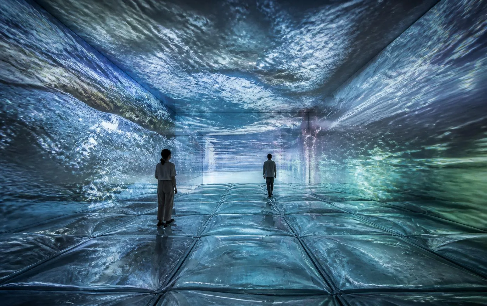 Walking on VateR — a full-room view in which documentary water footage and AI-generated water overlap continuously across the walls, ceiling and reflective water-filled floor, with two visitors present