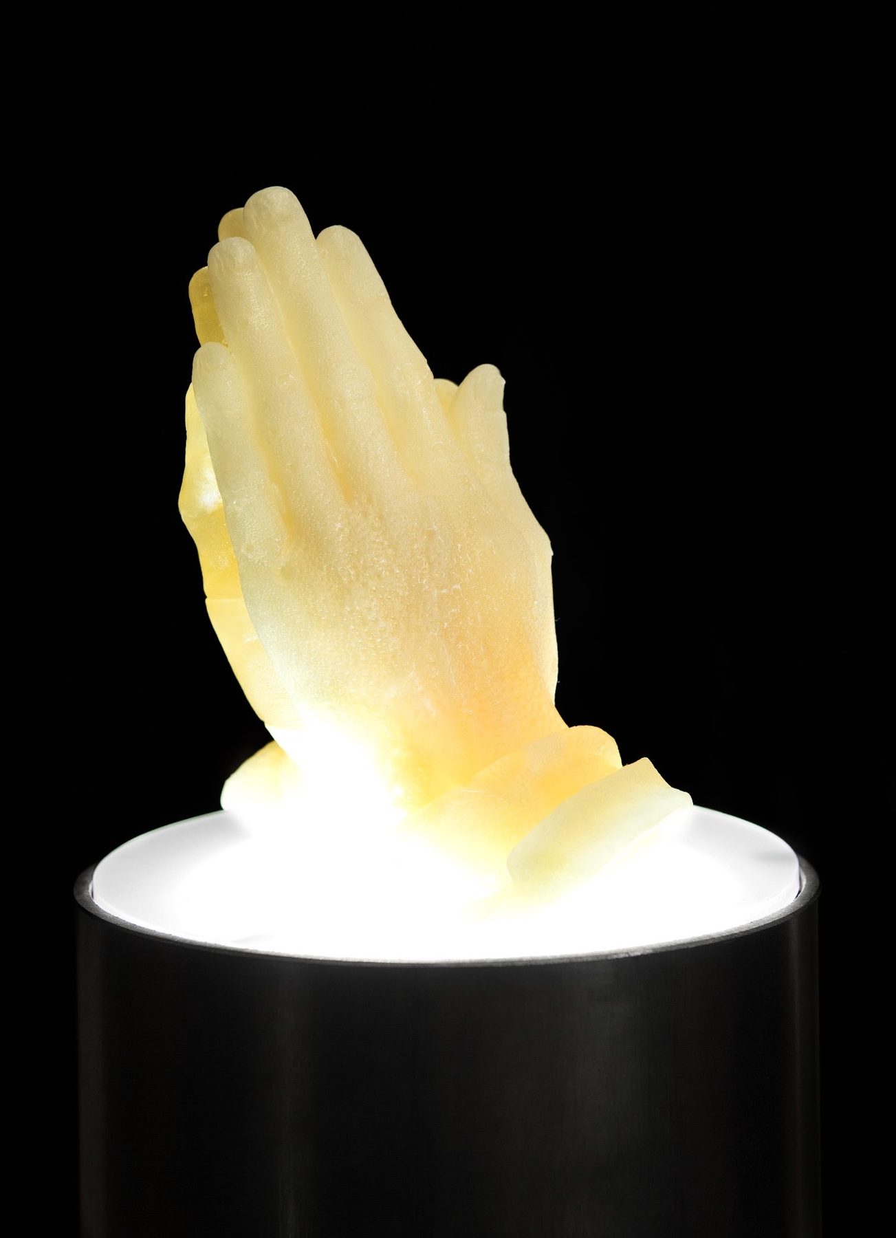 praYING hands, 2008 — hard-candy cast of praying hands in warm yellow, illuminated from below on a black pedestal, by Hannes Bend