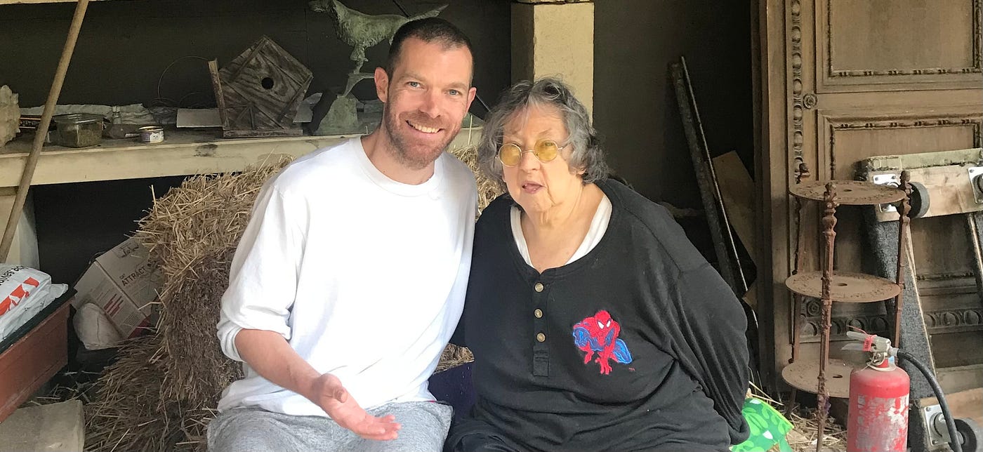 Hannes Bend sitting on a bale of straw in a barn next to an older woman with grey hair and yellow-tinted glasses; both are smiling at the camera.