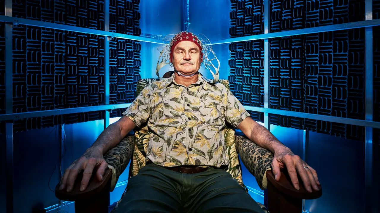 Joseph Kirschvink seated in an armchair inside a shielded chamber lined with dark acoustic foam, wearing a wired EEG cap, eyes closed.