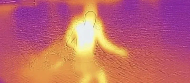 Thermal image of a human figure, with body heat glowing bright yellow and white against a purple-orange gradient background.