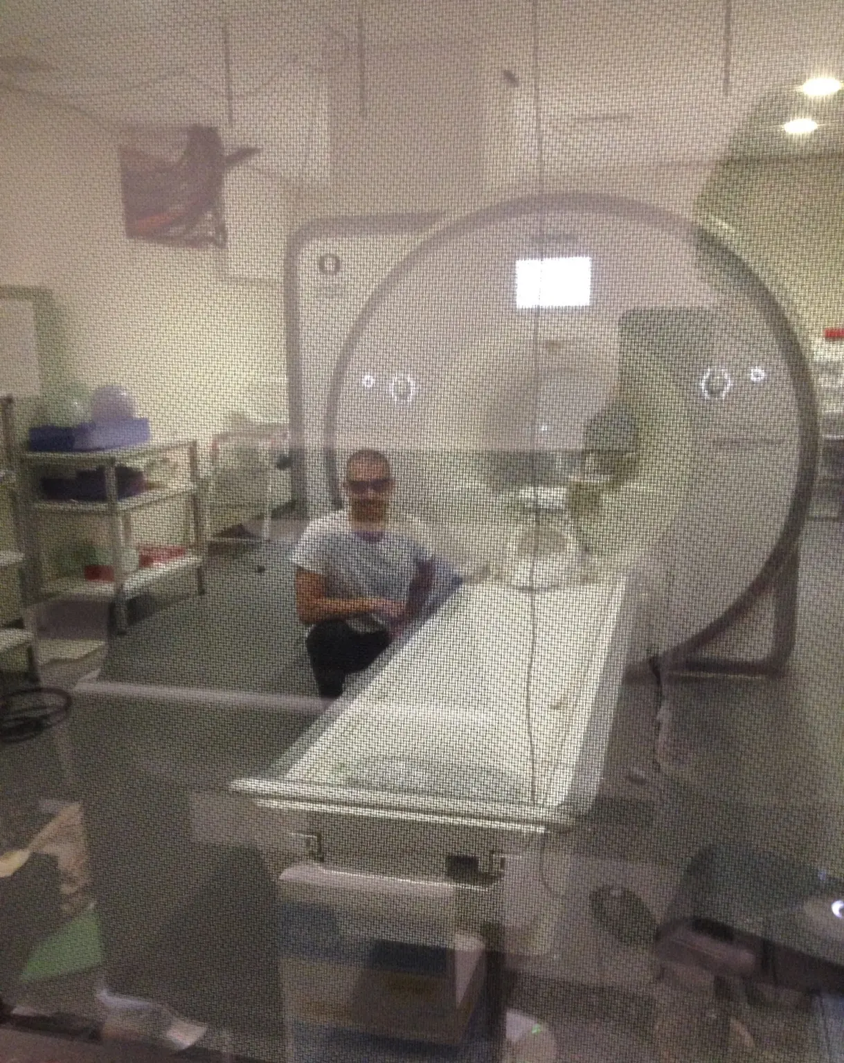 A person standing beside an MRI scanner and its patient table in a scanner room, seen through a control-room window.
