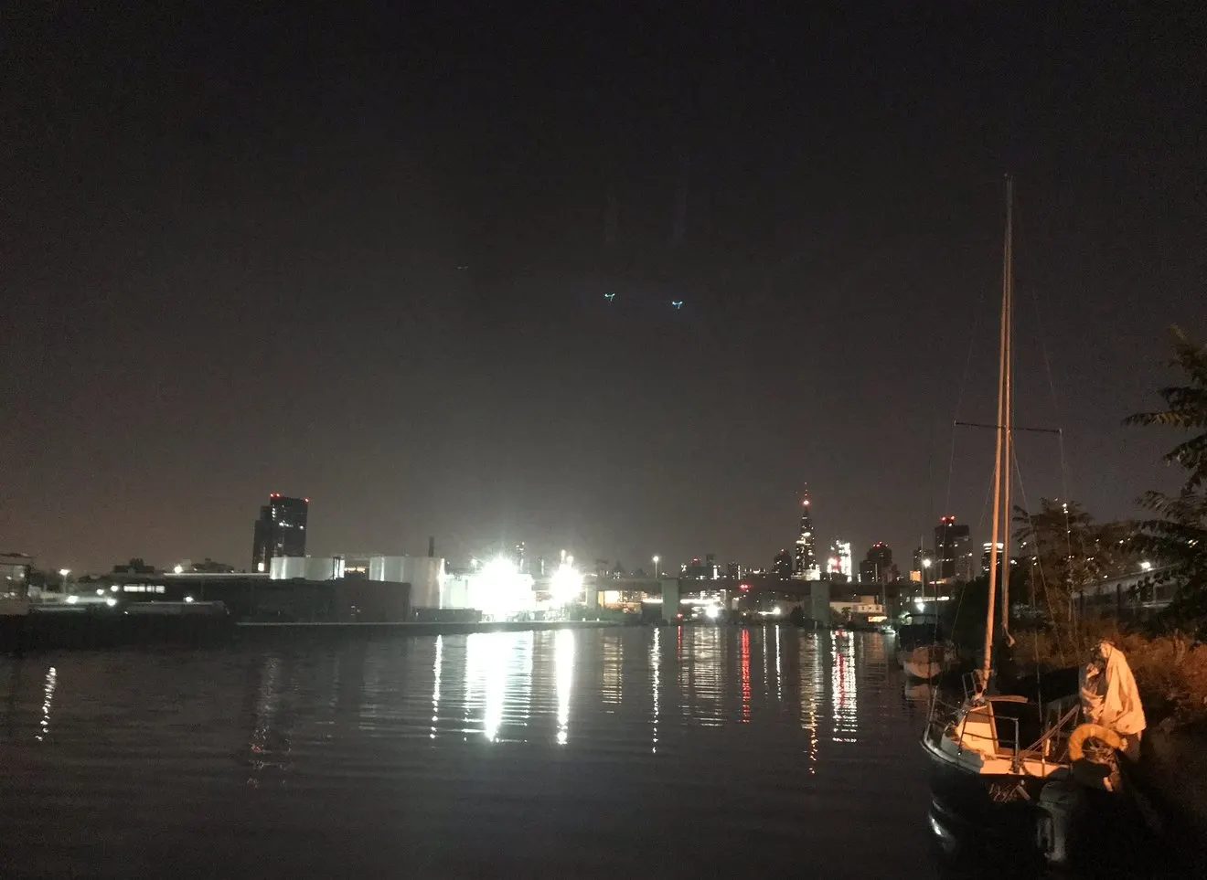 Hannes Bend's sailboat on Newtown Creek at night, with the New York skyline lit across the water.